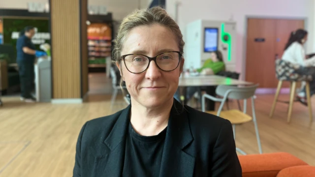Emily Huns sits for a photo in the University of Sussex's Student Life Centre lobby and communal area. She is wearing full-rimmed glasses, a t-shirt and a blazer, with her hair slicked back. Students in the background sit at tables studying.