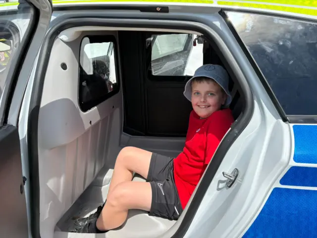 Bachgen yn eistedd yng ngefn car heddlu