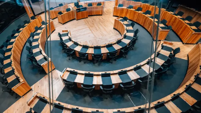 The Senedd chamber is a hemicircle of brown wooden desks with green chairs.
