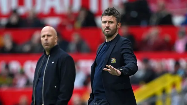 Carrick and Slot both dressed in black on the pitch sidelines