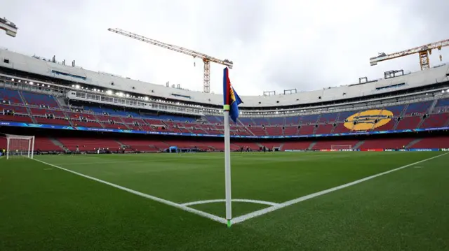 The Barcelona stadium, photographed from behind the corner flag (white stick with a red and blue flag) with a green pitch in the foreground and blue and red stadium seating wrapped around