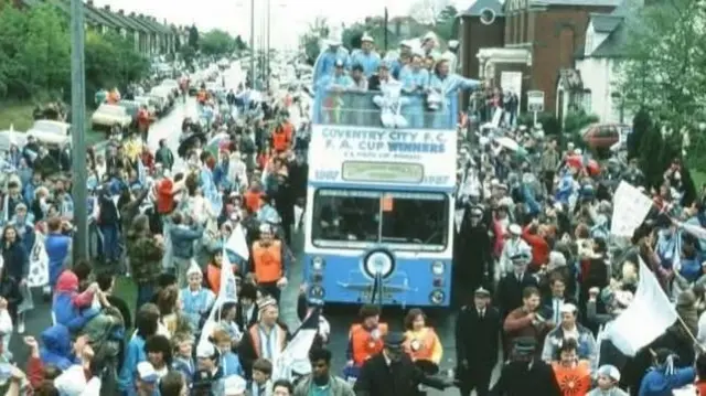 Crowds surround a blue bus which has FA Cup Winners written on it as it drives down a street