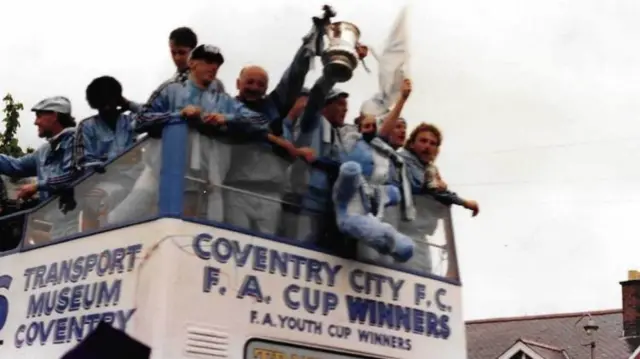 People at the front of a bus raise a trophy over the end and the bus has "Coventry City FC FA Cup Winners" on it