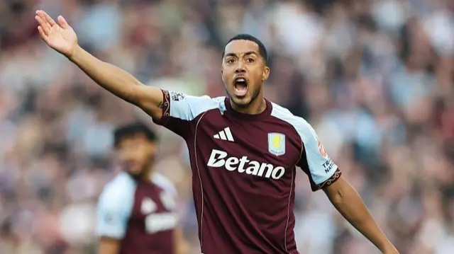 An Aston Villa player shouting with his arm up on the pitch. He is in a burgundy Villa top with light blue detailing.