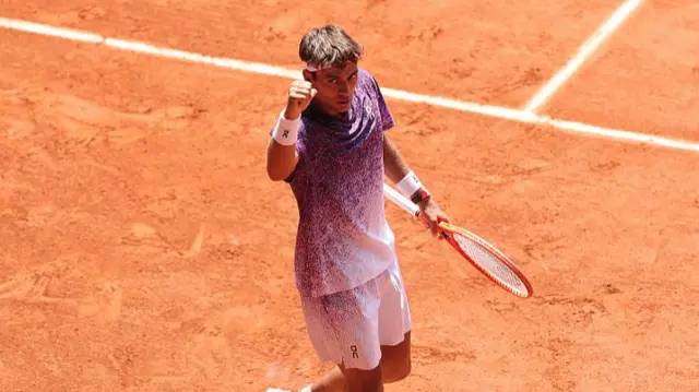 Flavio Cobolli, wearing a purple t-shirt and white shorts, holds his racquet in his left hand and raises his right fist
