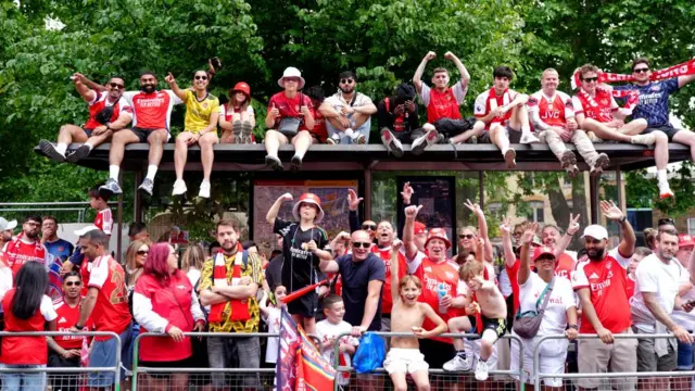 Fans stand on the pavement and sit on top of a bus stop to watch