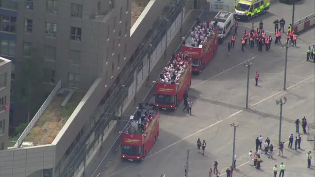 Three open-top red busses are parked with people standing on the upper deck of two of them