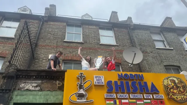 Three fans on a balcony overlooking the parade route hold up a replica trophy as they cheer towards the photographer