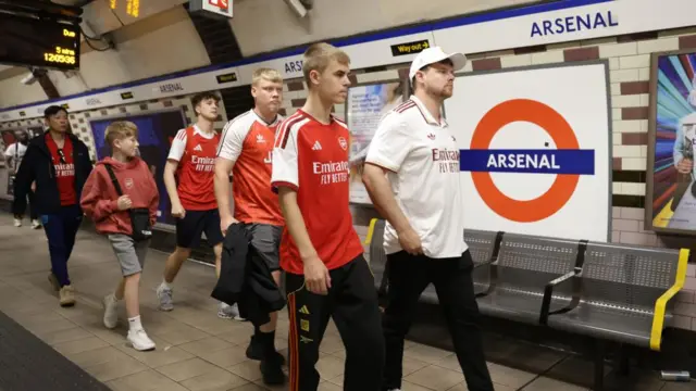 Group of Arsenal supporters walking along platform at Arsenal station, wearing red and white shirts