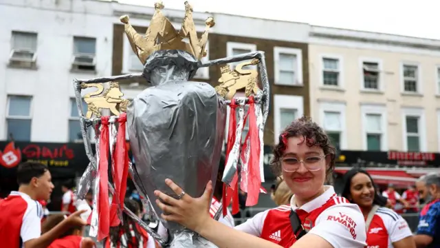 A woman holds a paper mache trophy on the street among Arsenal fans