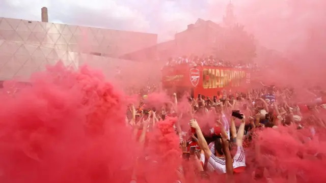 Red smoke fills the air as the buses pass large crowds
