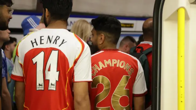 Two people wearing Arsenal jerseys, “Henry 14” and “Champions 26,” on crowded underground platform