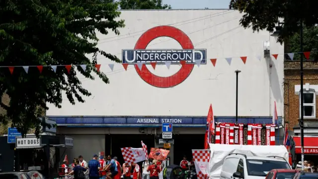 A shot of the entrance to Arsenal Underground Station. It is decorated with Arsenal FC flags and red and white bunting.