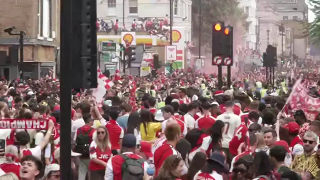 Fans line the streets, some stand on top of a garage to get a good view