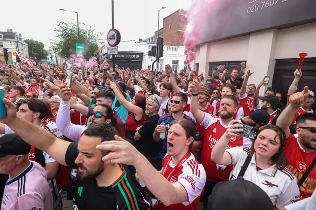 Fans cheer as a bus goes past and red flares go off in the distance