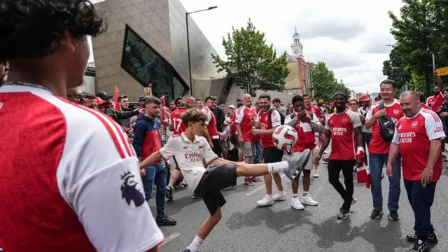 A young arsenal fan does kicky uppies on the road as other fans look on