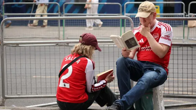A man and woman sit on the floor and read books behind silver railings ready for the parade