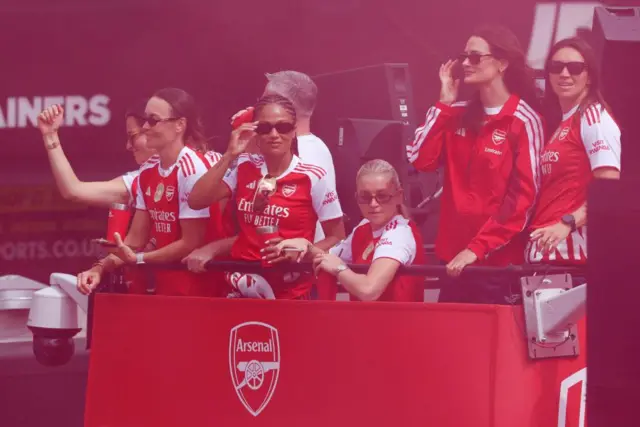 Players in Arsenal kits standing on open-top bus, surrounded by pinkish red flare