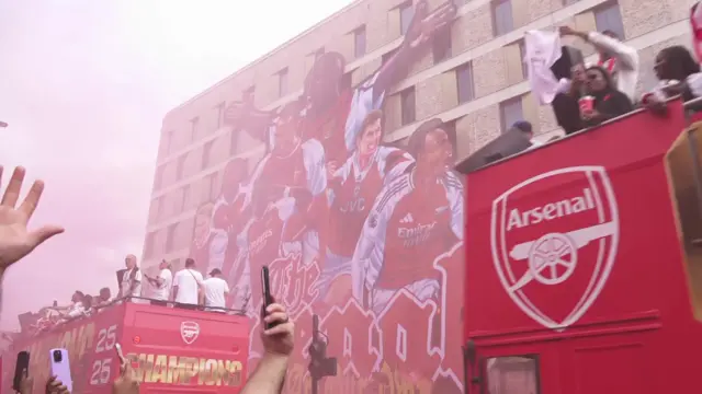 The buses pass by while a huge Arsenal banner covers a building behind