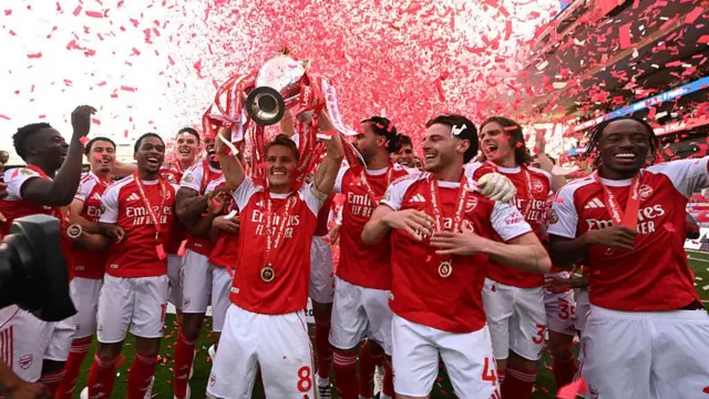 Arsenal's captain Martin Ødegaard surrounded by team members lifting the trophy as red confetti rains on them
