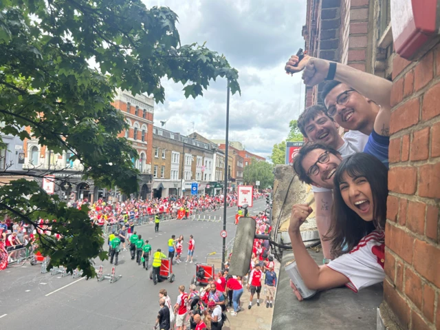 To the left: a street in Islington with Arsenal fans crowding on both sides, as officials stand in the middle. To the right, emerging from a flat window, are three young men and a woman cheering and smiling