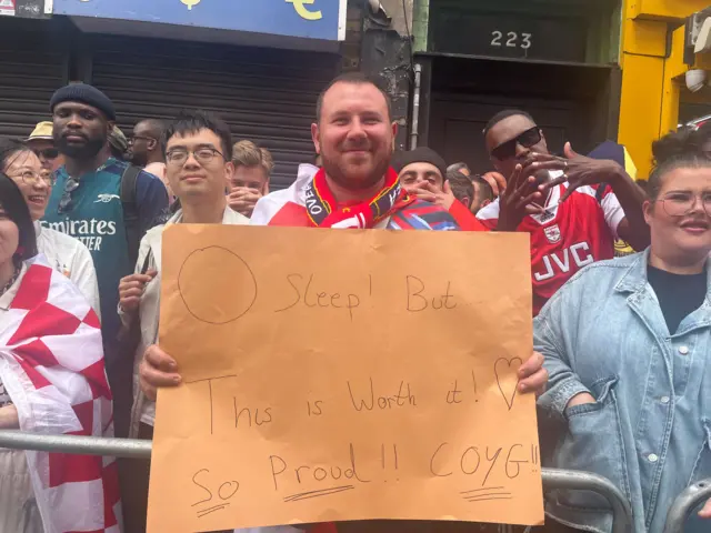 Young man in Arsenal shirt with a team scarf around his neck smiles as he holds up a sign on orange paper reading "0 sleep! But... this is worth it (heart drawing) So Proud!! (underlined) COYG!! (underlined)"