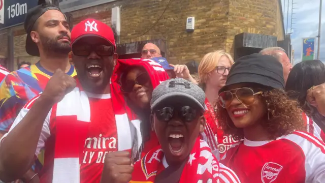 From left to right: A black man in Arsenal kit and a red New York Yankees hat cheers as his teenage daughter, in red Arsenal hoodie and black sunglasses, leans on his left shoulder. A young black boy, also in full Arsenal kit (red shirt with white and red stripes on shoulders) and a grey hat, cheers in the foreground as his mum (in black bucket hat, Arsenal shirt and aviator sunglasses) smiles next to him
