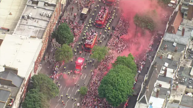 Buses seen from the air - red smoke comes from the large crowd either side