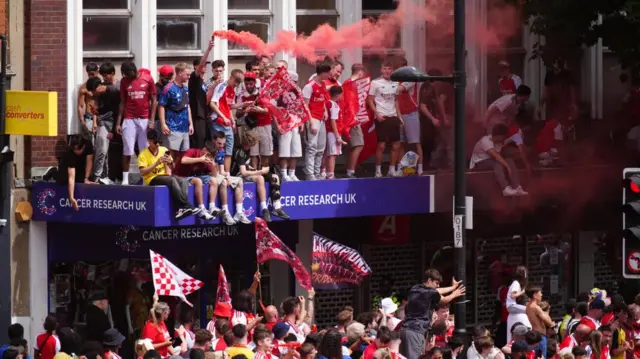 Fans stand on top of the entrance above a shop and hold flares and flags