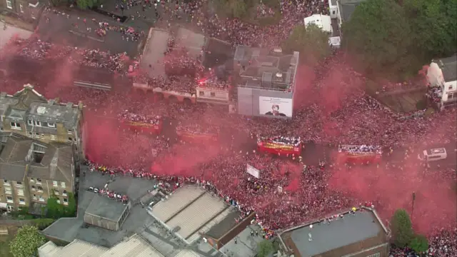 The parade as seen from the air - people stand on rooftops and red smokes fill the air