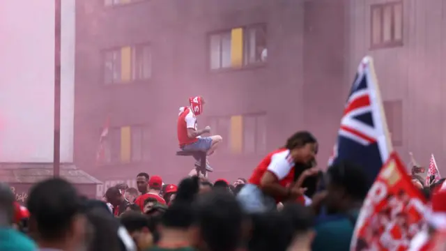 A man sits on a lamppost amid a sea of fans