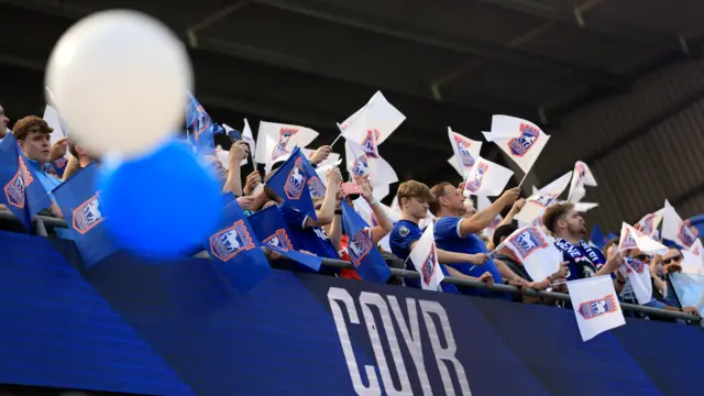 Ipswich Town fans in the stands at Portman Road stadium on Saturday. Fans are waving blue and white flags in the air.