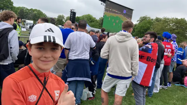 A boy in an orange Ipswich Town shirt poses with a thumb up against the backdrop of fans gathered in Christchurch Park