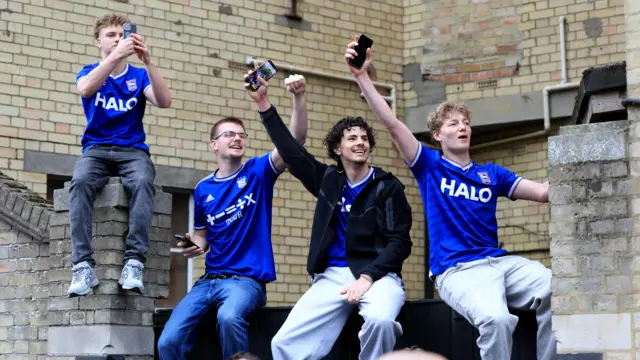 A group of four boys in Ipswich Town football shirts trying to get a glimpse of the parade bus. They are all smiling and are capturing the moment on their phones