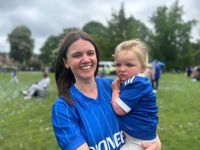 A woman with shoulder length brown hair holds a small girl. Both have blue Ipswich Town shirts on