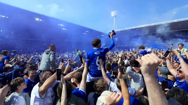 Fans take part in a pitch invasion after Ipswich Town is promoted. There are hundreds of people on the pitch, some people carrying fans on their shoulders. Flares have been let off and people are capturing the moment on their phones.