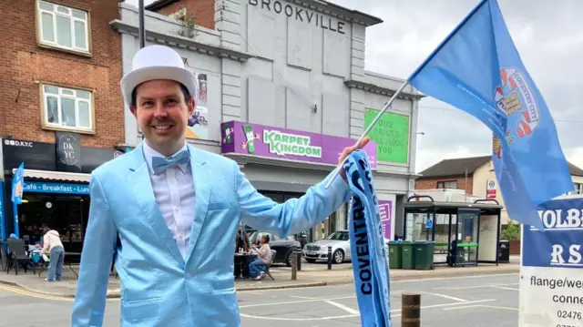 A man in a top hat, wearing a sky blue jacket, is waving a Coventry flag
