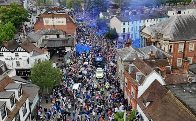 Aerial view of hundreds of people lining the route of an open top bus parade through Ipswich