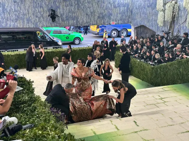A group of people help a woman in a voluminous dress up some stairs