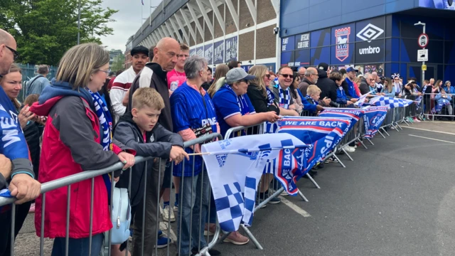 Crowds of people line metal barriers at Portman Road Stadium.