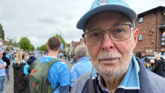A man with a short white beard and wearing black-rimmed glasses looks into the camera while wearing a blue baseball cap