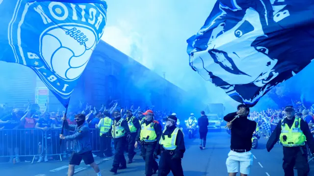 Ipswich Town fans line the street inside metal barriers awaiting the players arrival ahead of the match. Blue flares have been let off and police officers show the way.