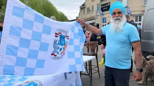 A man with a white beard and sky blue turban holds up a Coventry flag