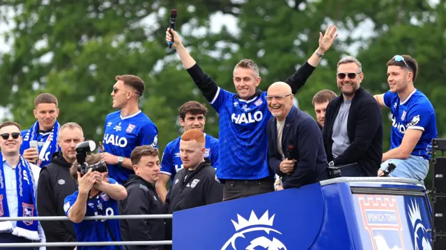 Kieran McKenna on the top of the open top Ipswich Town bus during the parade. He is wearing a blue Ipswich Town football shirt and has his arms in the air.