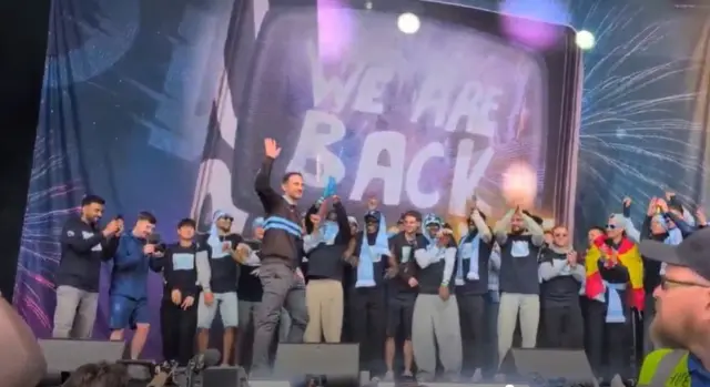 A group of men waving their hands on a stage in front of a sign saying "We are Back