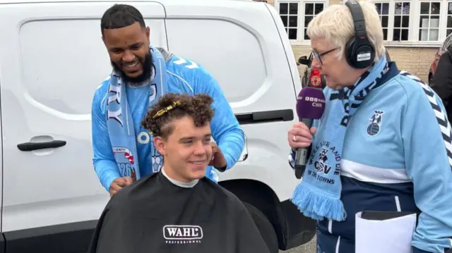 A teenager with a hair dressers cape around him gets a hair cut from a man wearing a sky blues scarf