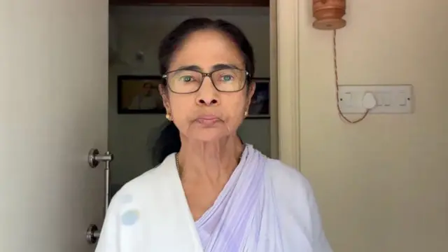 A mid close up of Mamata Banerjee who is wearing a white saree with a purple background