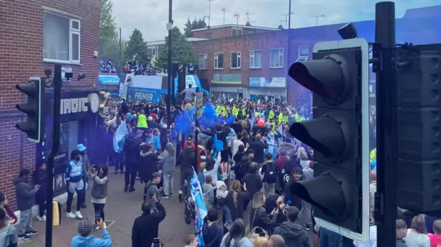 Crowds around the bus as it slows through a street.