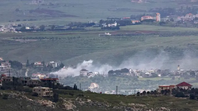 Wide view of rural hills shows homes and farms, with white smoke drifting above a village amid green fields landscape