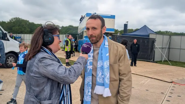 Liam Kelly wearing a beige jacket and a light blue Coventry City scarf is interviewed by a BBC CWR reporter with a purple microphone.